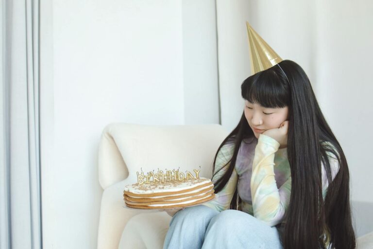 A woman in a party hat looking at a birthday cake with a thoughtful expression indoors.