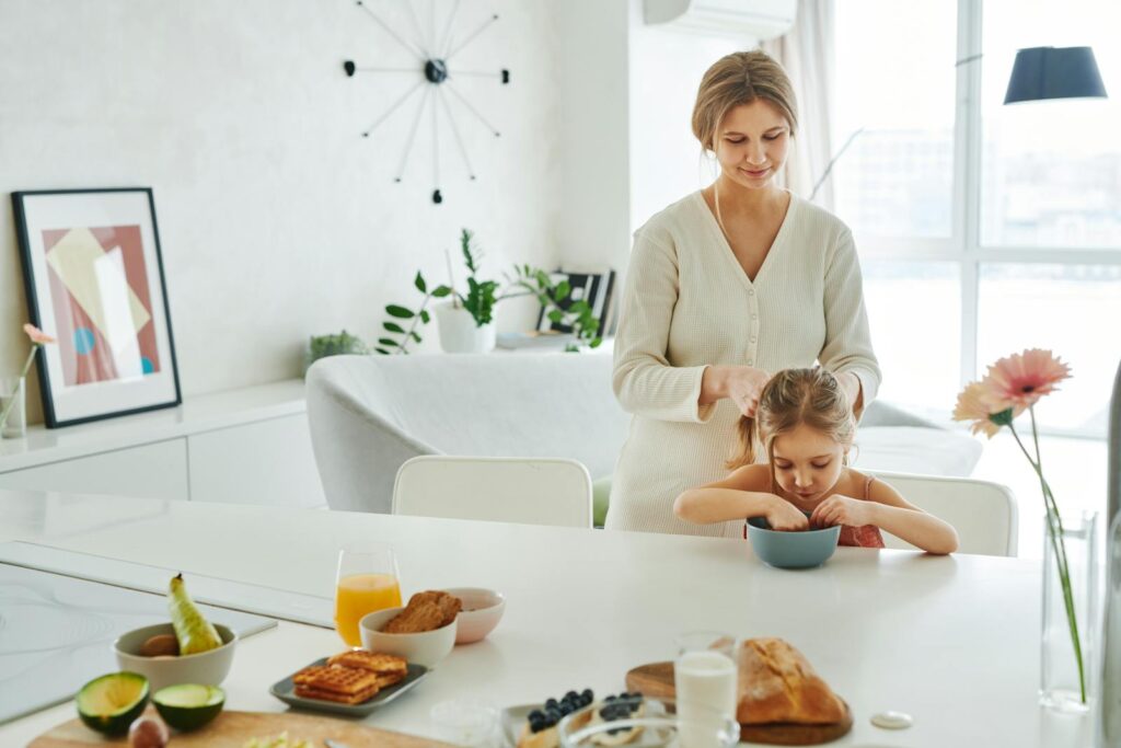 A mother braids her daughter's hair in a bright kitchen, surrounded by breakfast items.