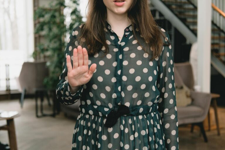 Young woman in polka dot dress gesturing stop with hand indoors, displaying confident attitude.