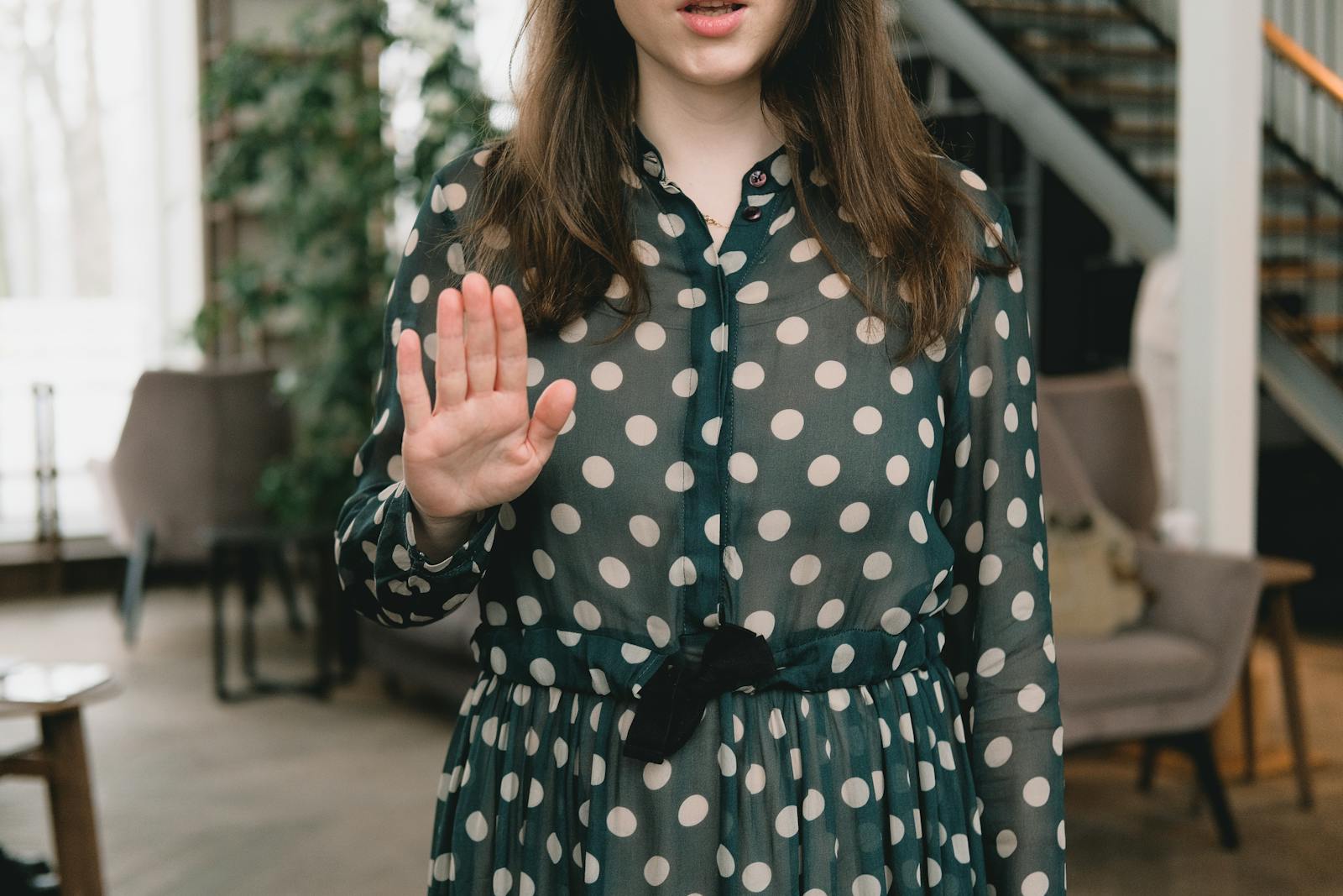 Young woman in polka dot dress gesturing stop with hand indoors, displaying confident attitude.