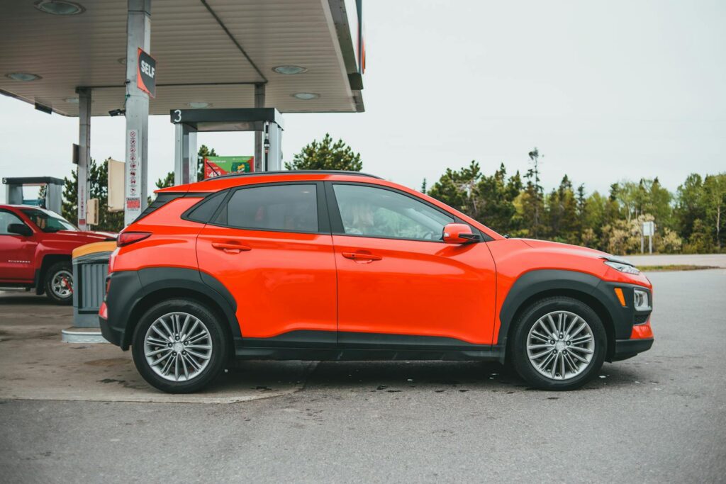 Bright orange SUV parked at a gasoline station with a red truck in the background.
