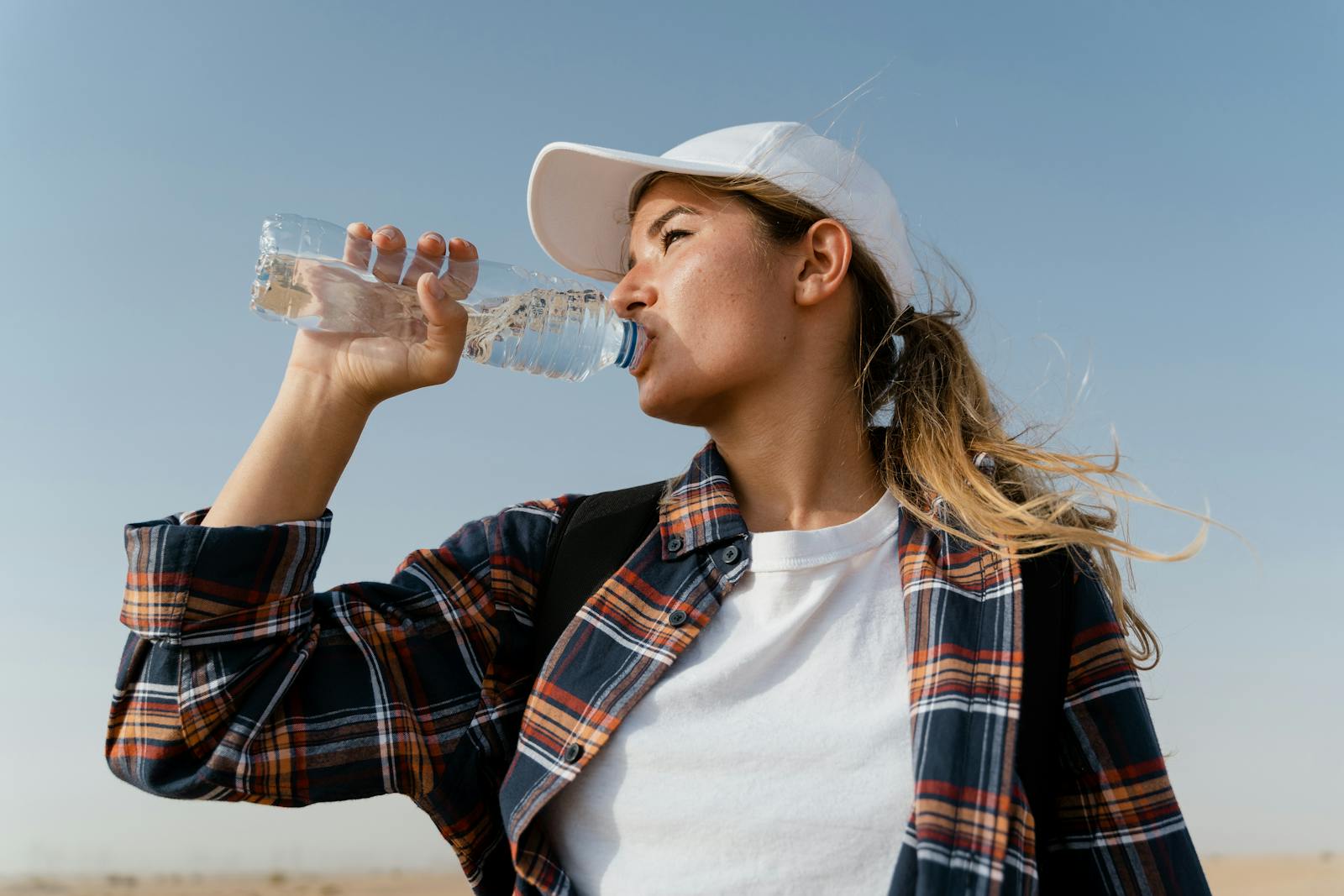 A woman in a baseball cap drinks water from a bottle under a clear blue sky.