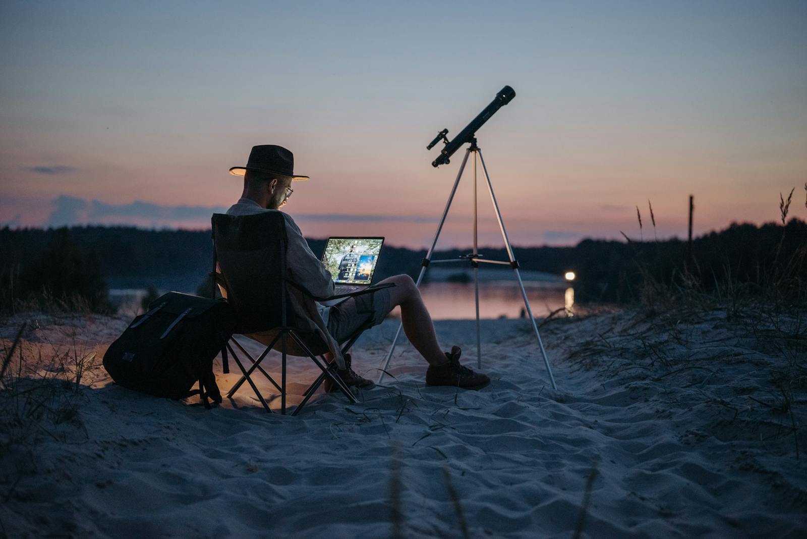 A man works on a laptop with a telescope nearby, on a sandy beach at twilight.