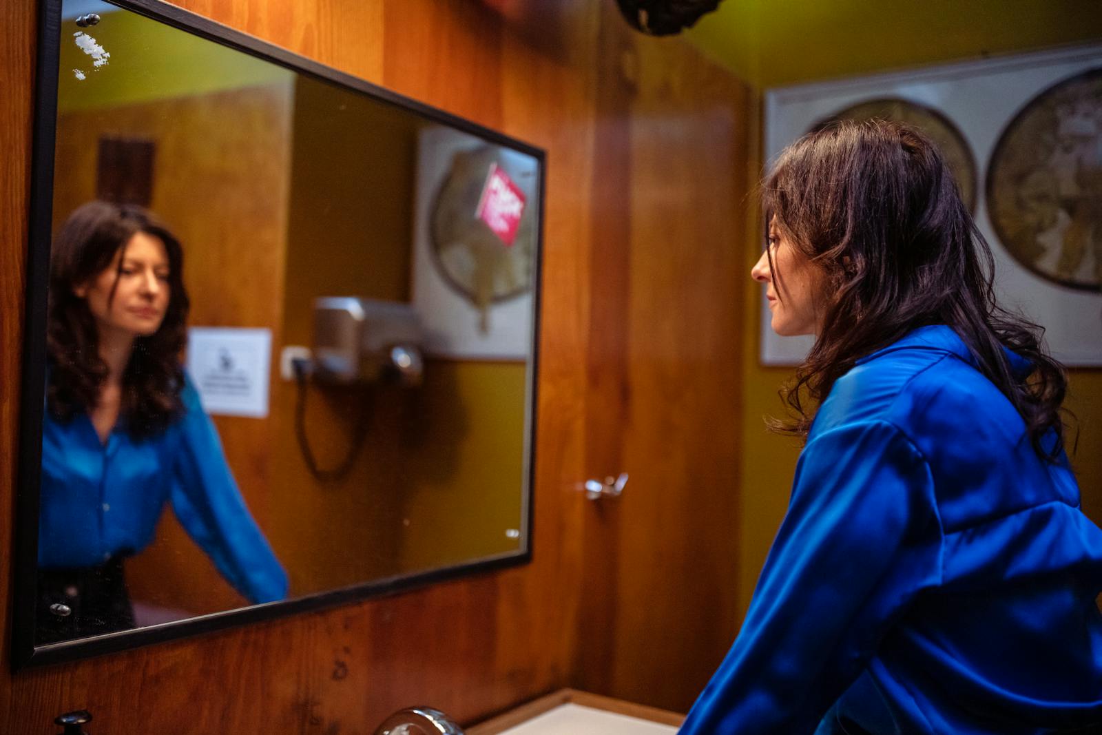 Woman in blue shirt reflected in restroom mirror with wood paneling. Indoor scene with warm tones.