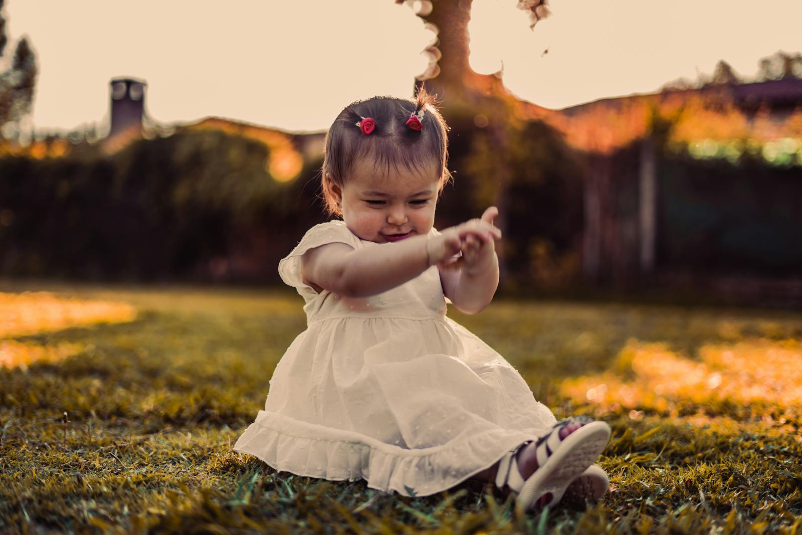 Adorable baby girl in white dress playing on grass during sunset, capturing joy and innocence.