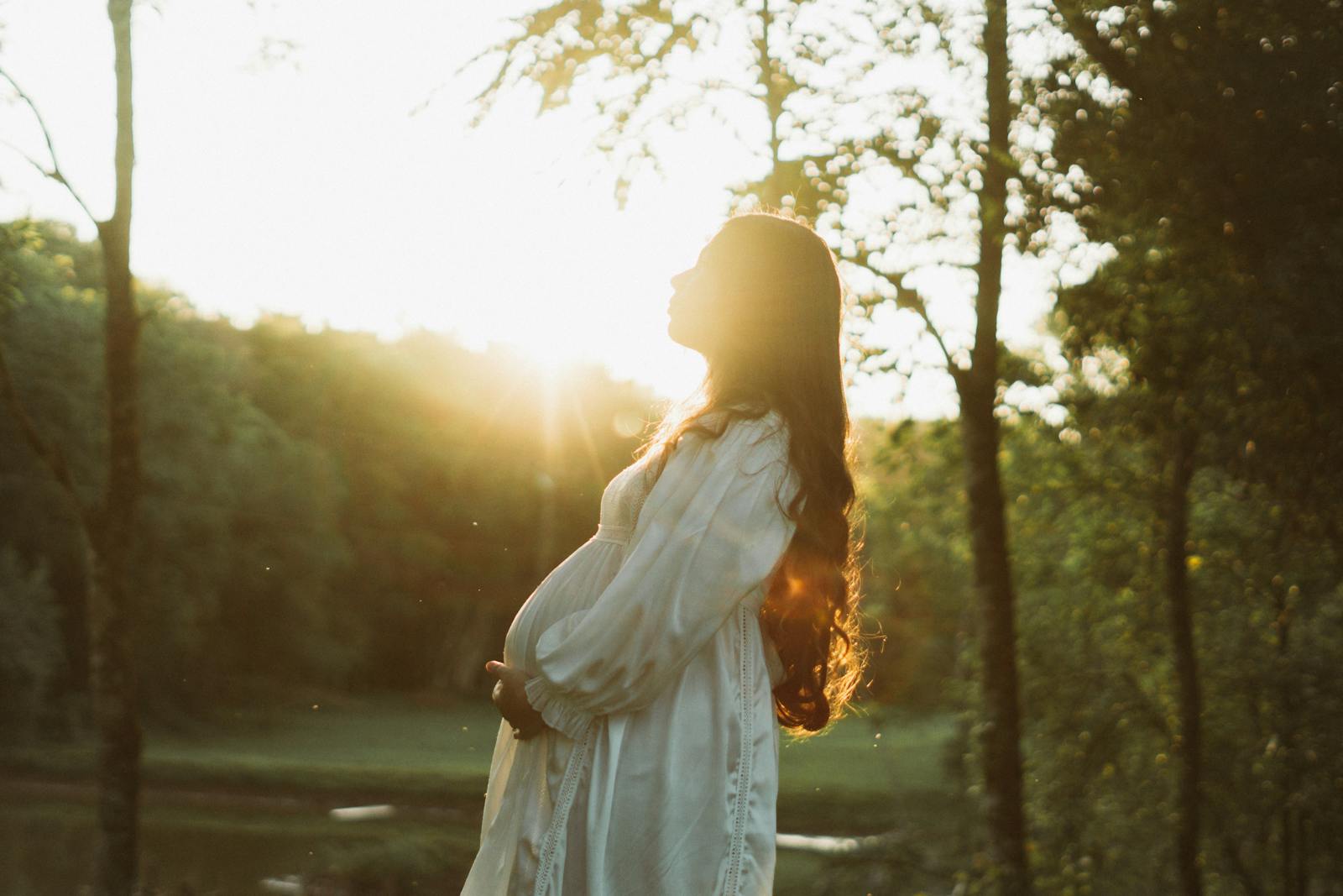 Pregnant woman in white dress enjoying a sunset in a serene forest setting.