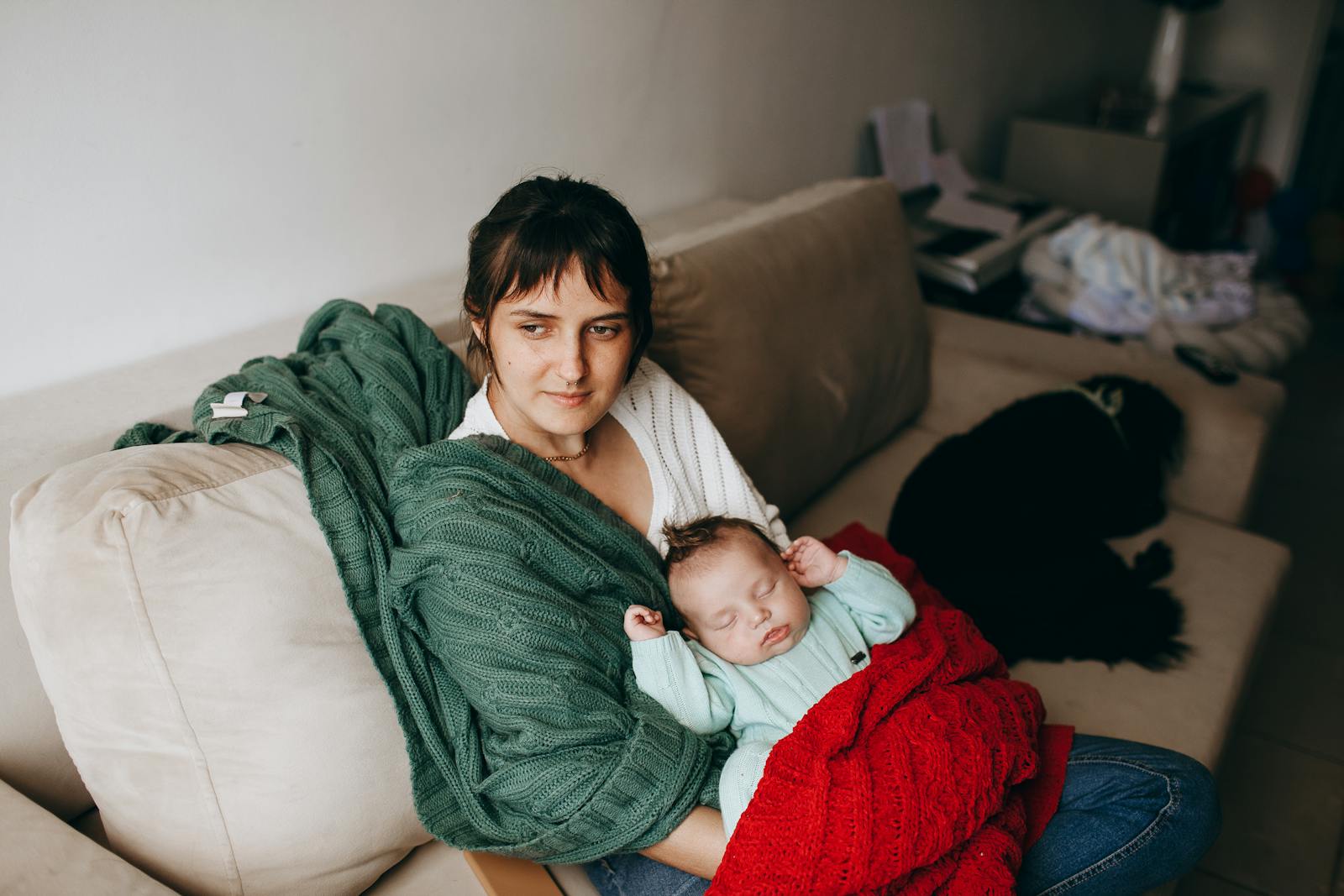 A mother lovingly holds her sleeping baby on a cozy sofa, surrounded by warm blankets.