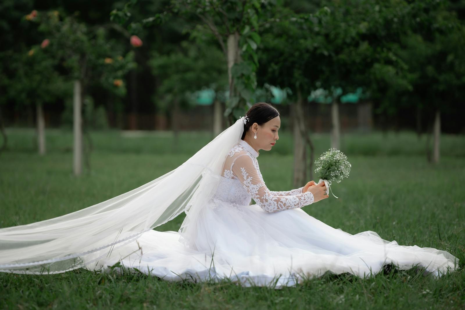 Elegant bride in a white wedding dress sitting on grass holding bouquet in a lush garden.