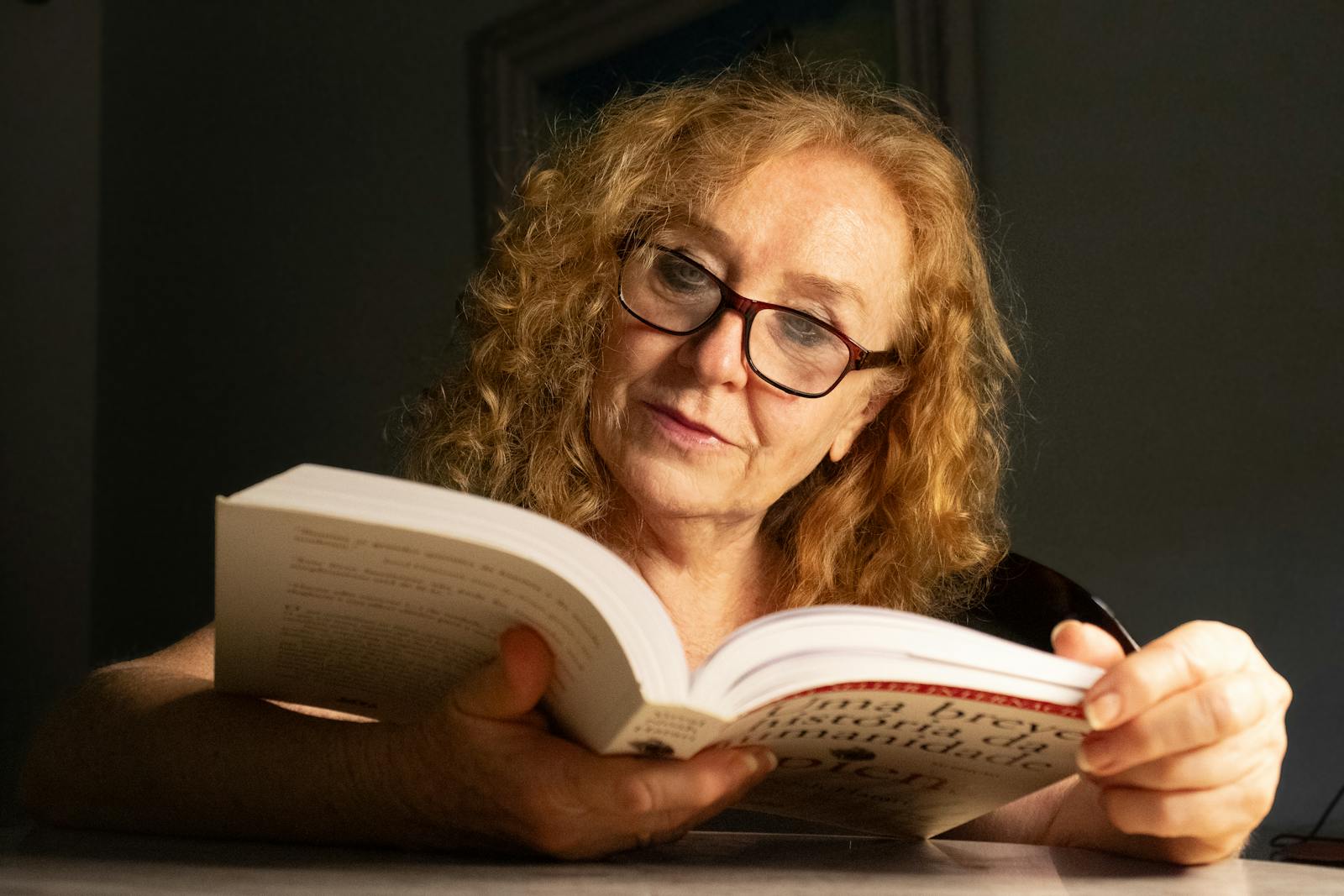 Senior woman with glasses reading a book in a cozy indoor setting, focused and relaxed.