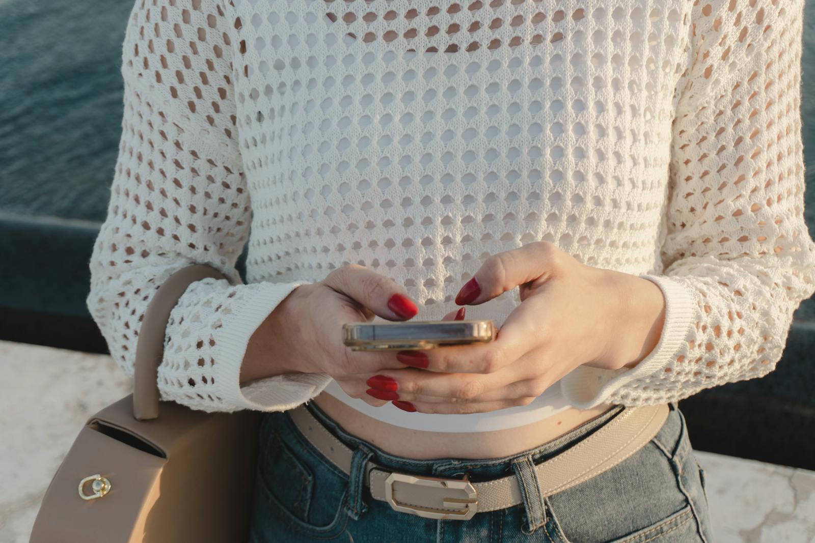 Close-up of a woman using a smartphone outdoors, wearing a white knitted top and jeans.