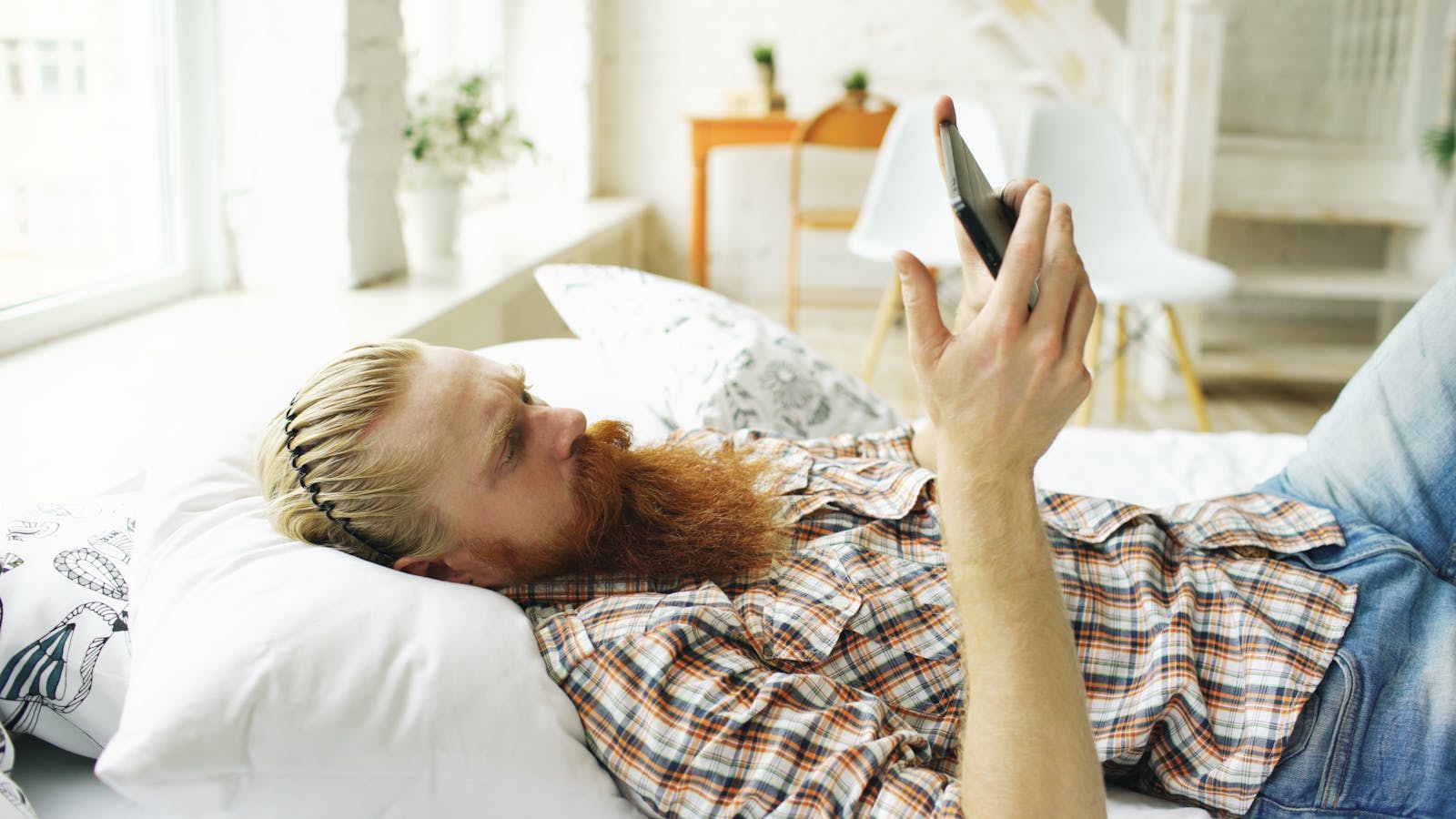 Casual man with a beard relaxing on bed, using smartphone in bright room.