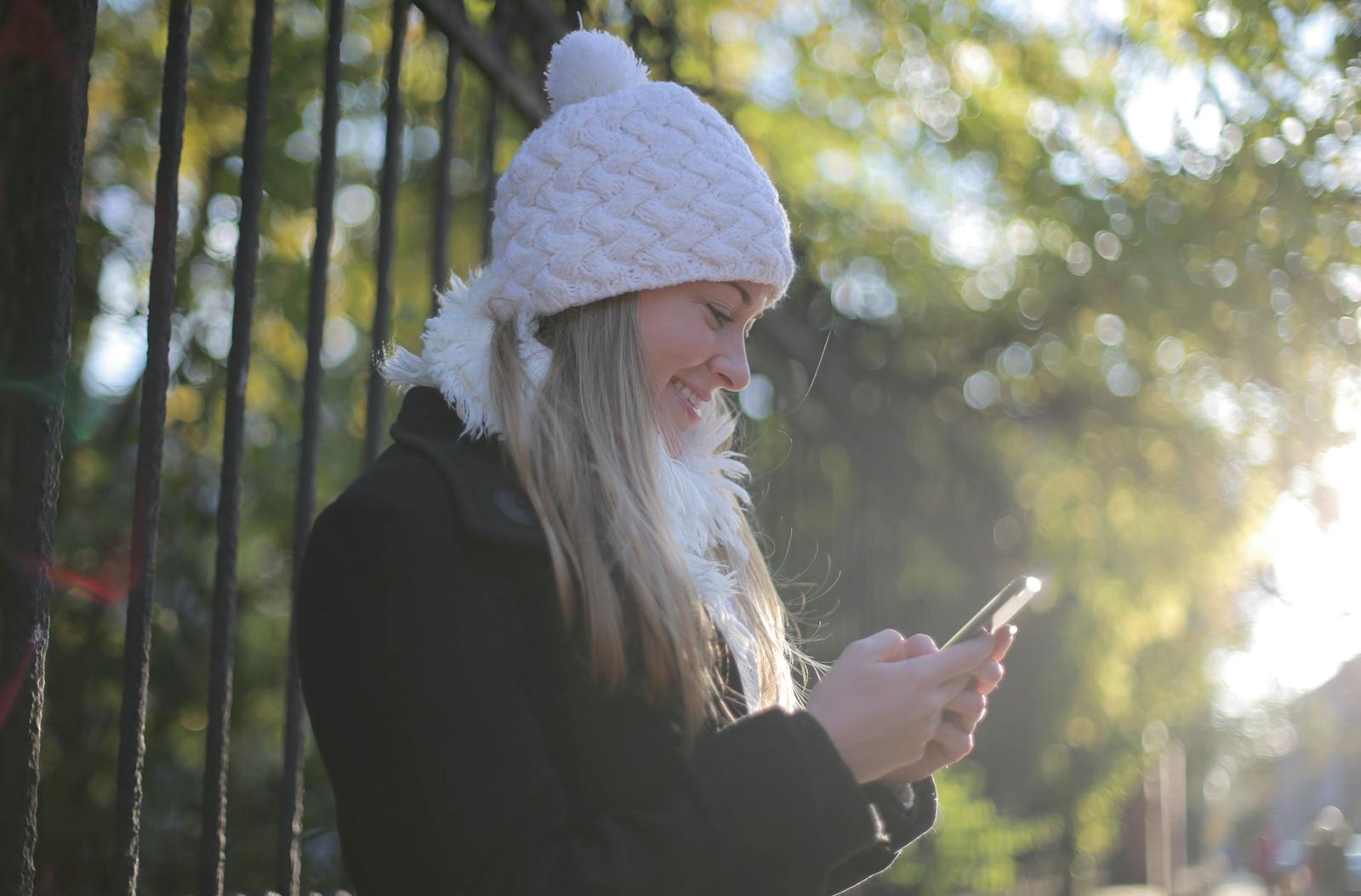 A smiling woman in a white beanie texting on her phone outdoors during winter.