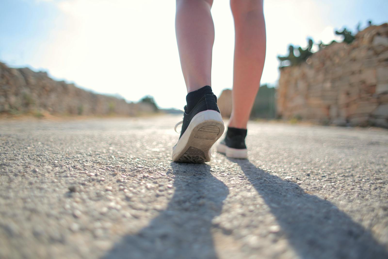 A person walks on a sunny rural road wearing sneakers, casting a long shadow.