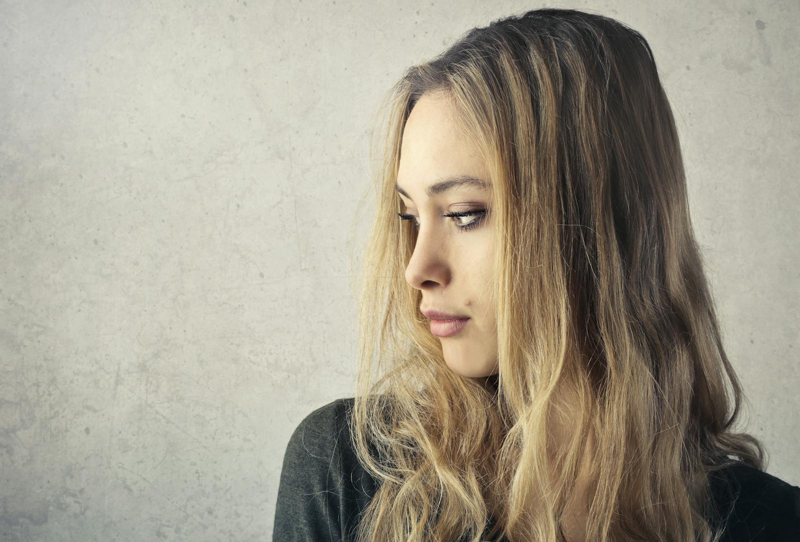 A pensive young woman with long blonde hair looking to the side against a neutral background.