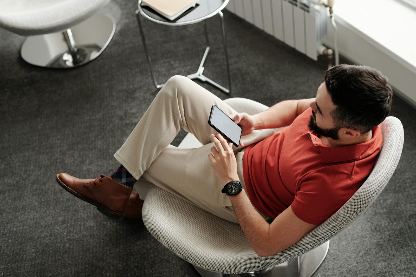 Bearded man in a red polo shirt sitting comfortably, using a smartphone indoors.