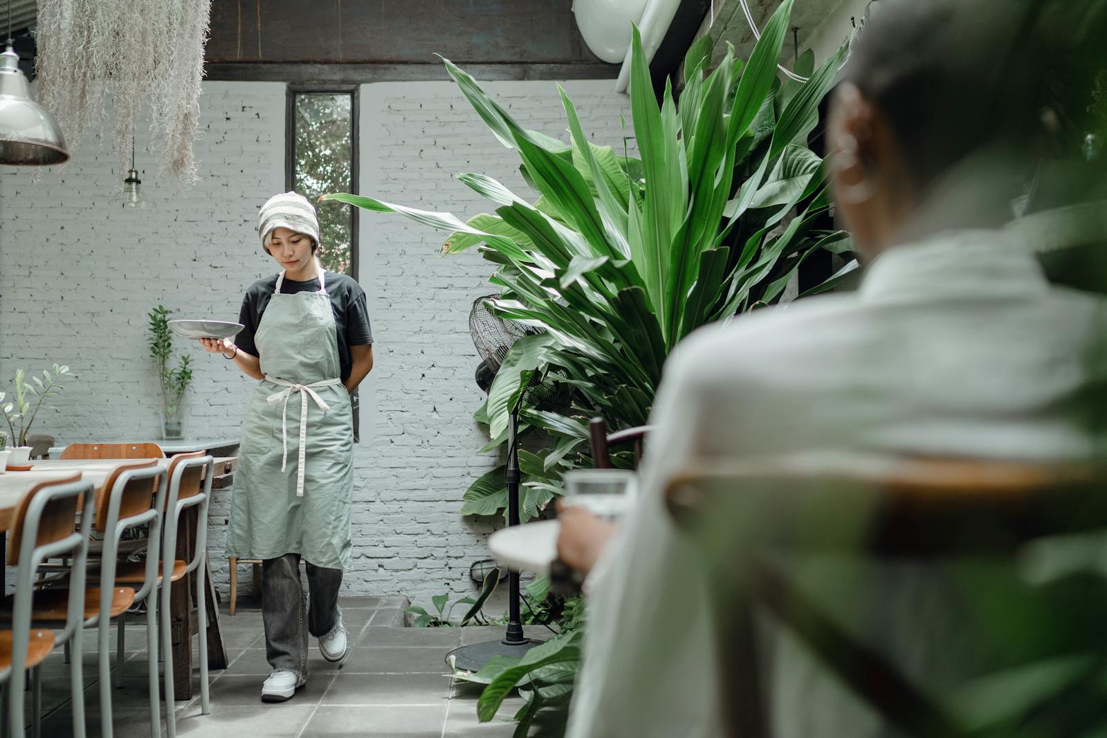 Positive young waitress in hat and apron with plate walking along tables in creative cozy restaurant towards guest sitting near big green plate