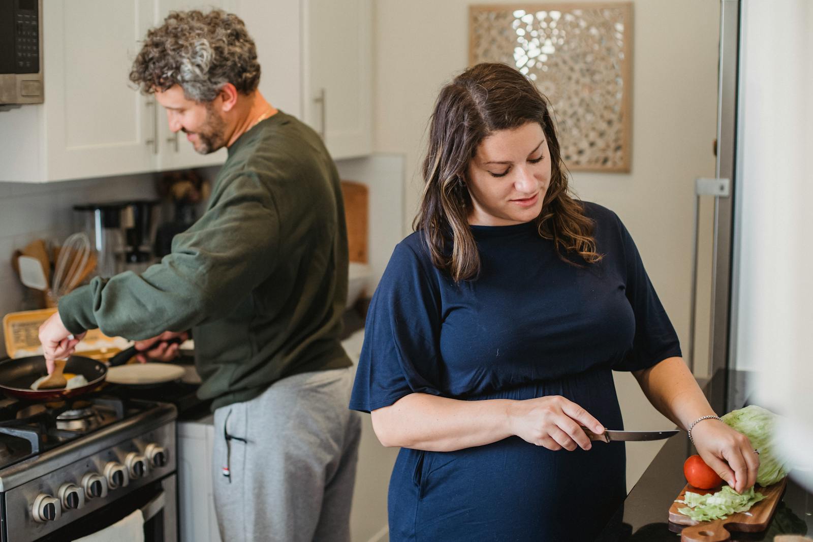 Adult pregnant woman in casual dress cutting vegetables while husband cooking meal on heater in contemporary light kitchen