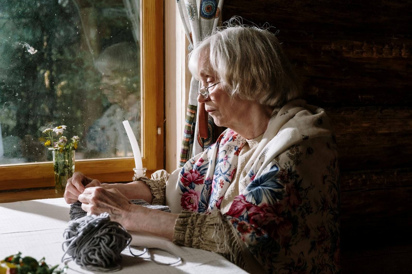 Senior woman knitting by a sunlit window in cozy interior, embracing a peaceful hobby.