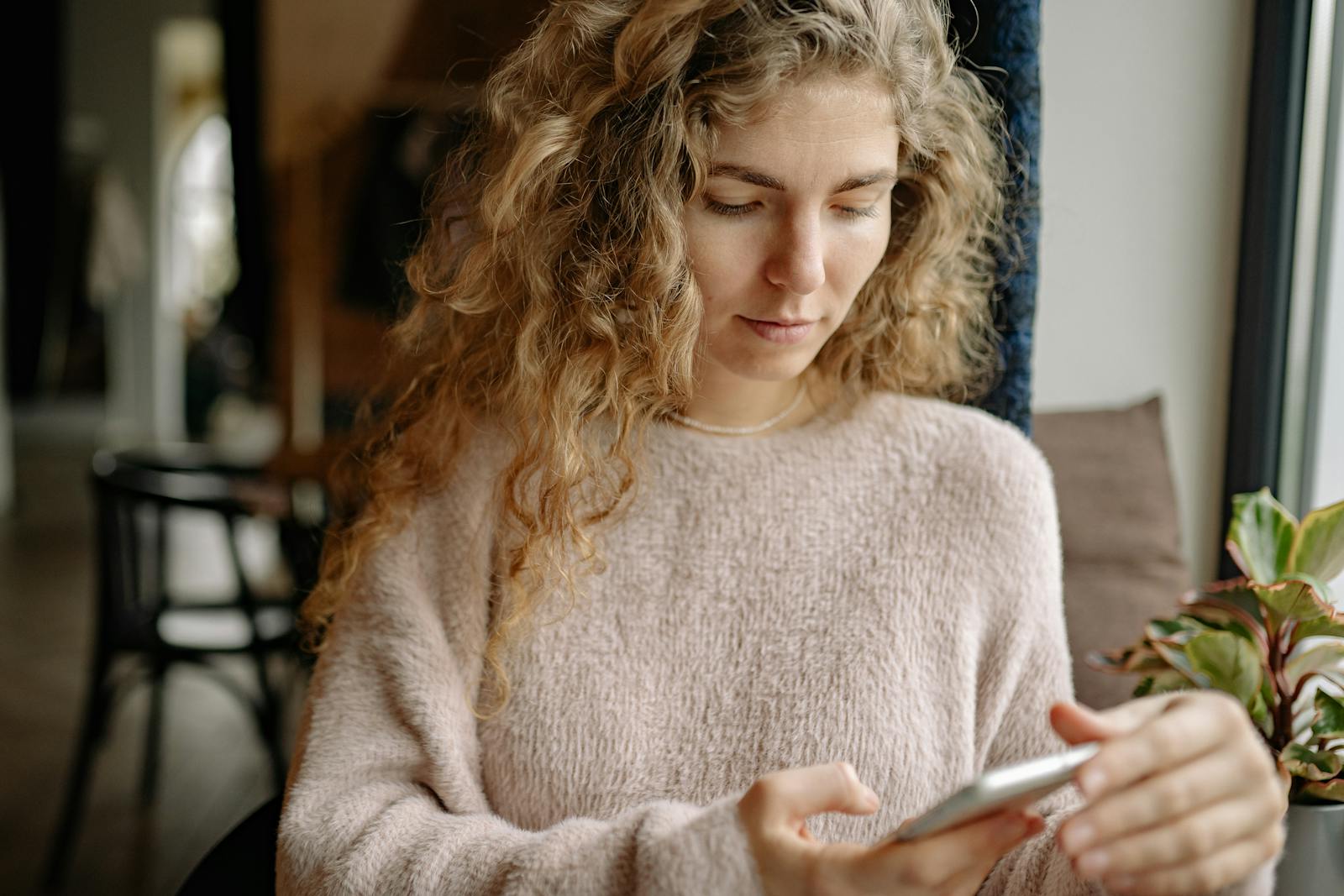A woman in a cozy sweater using a smartphone in a relaxed indoor environment.