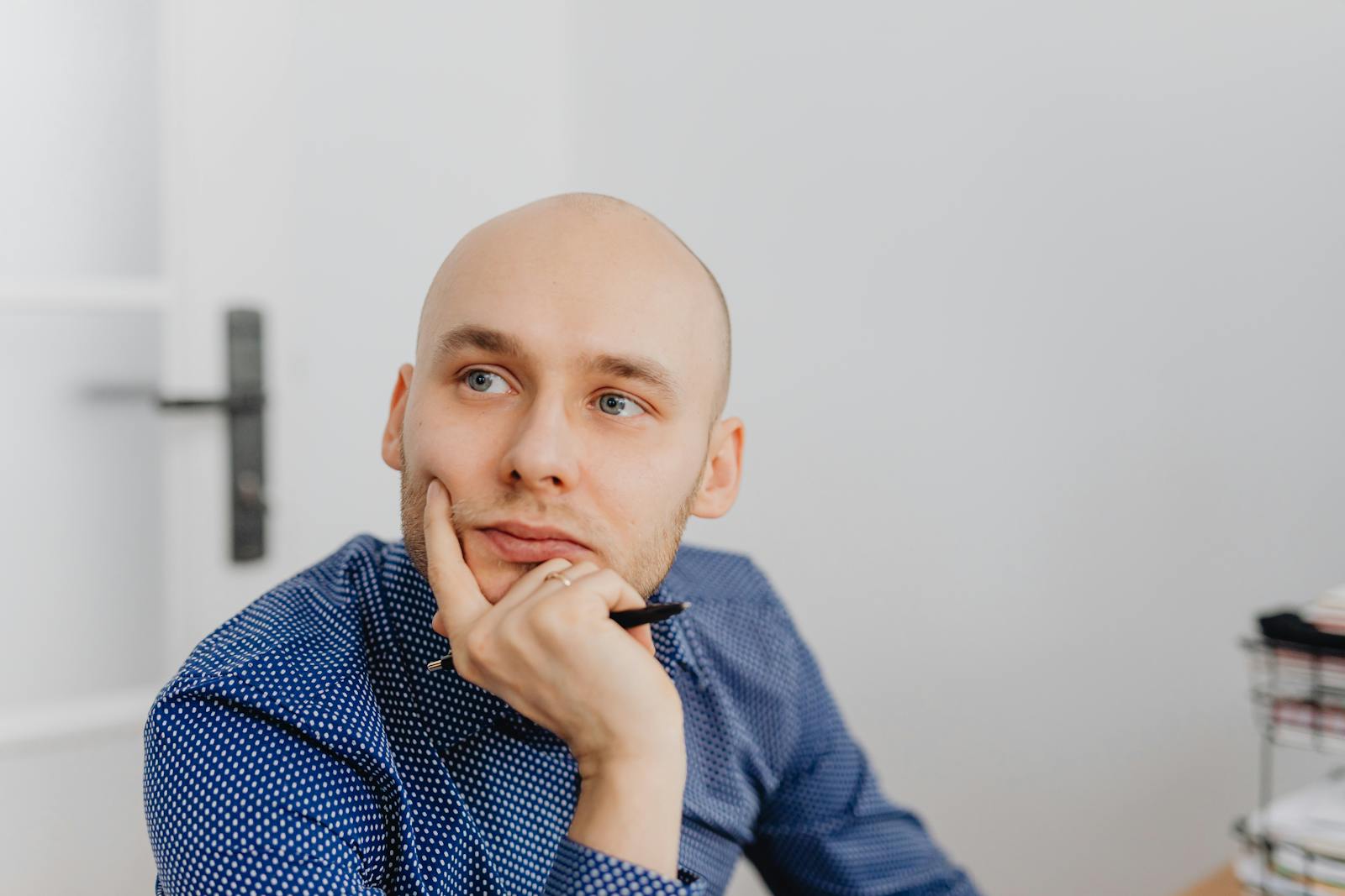 Bald man in blue shirt thoughtfully holding a pen indoors, gazing into distance.