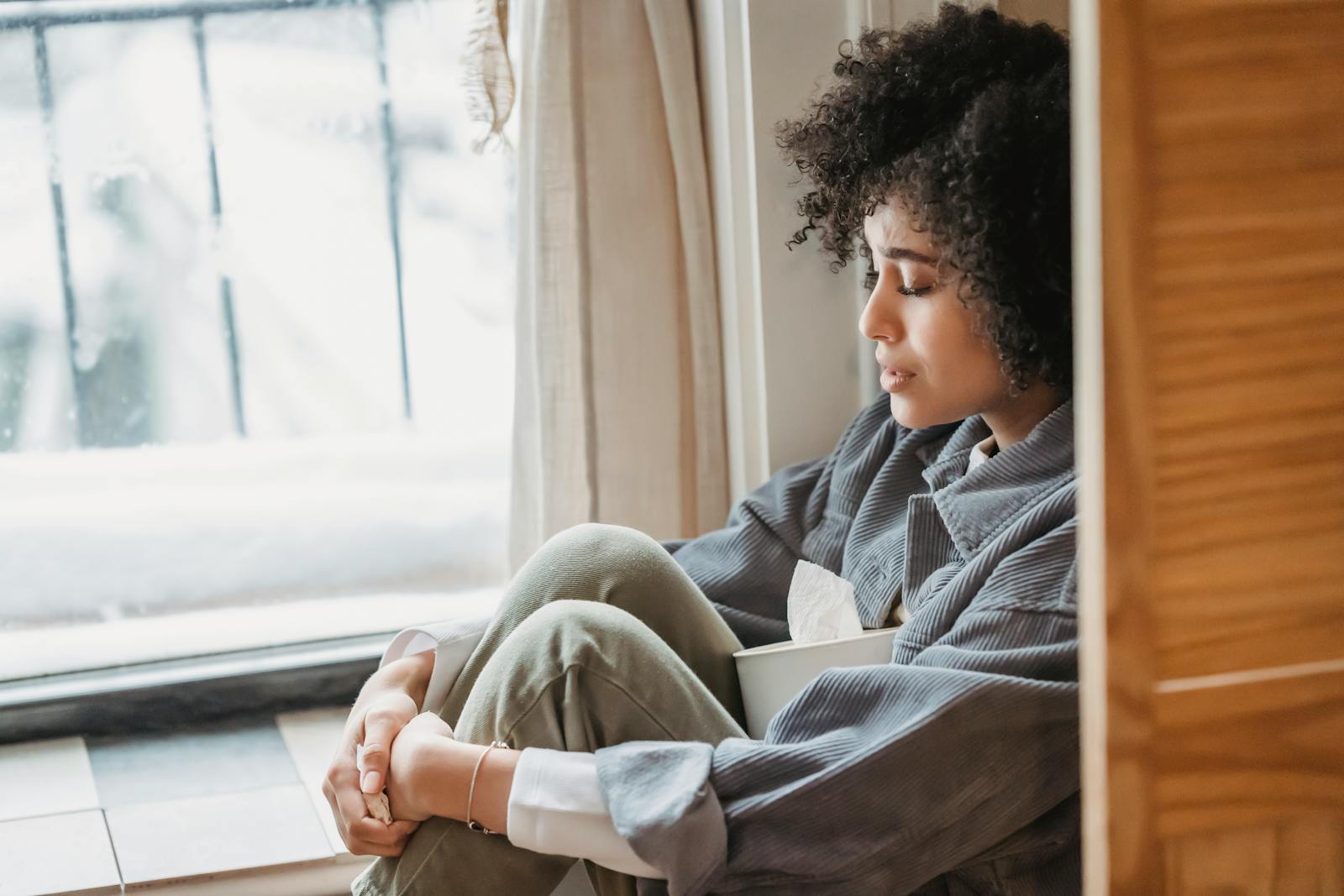 High angle side view of crying African American female with curly hair sitting on windowsill with box of tissues