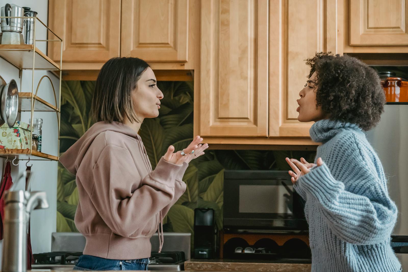 Side view of unhappy young multiracial girlfriends in casual clothes standing in kitchen while having disagreement and looking at each other