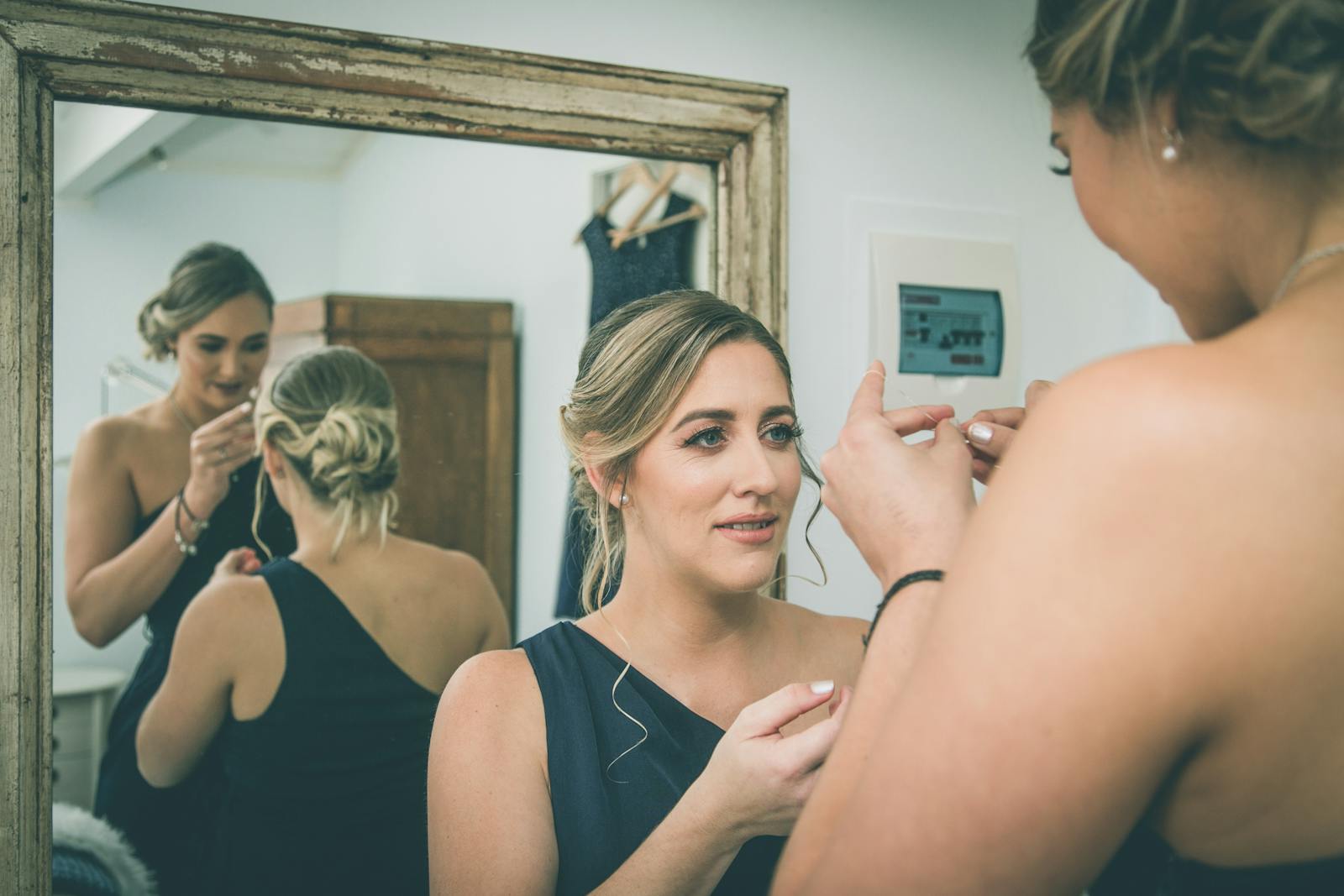 Stylish young female friends in elegant similar dresses standing near window in room and preparing for wedding party
