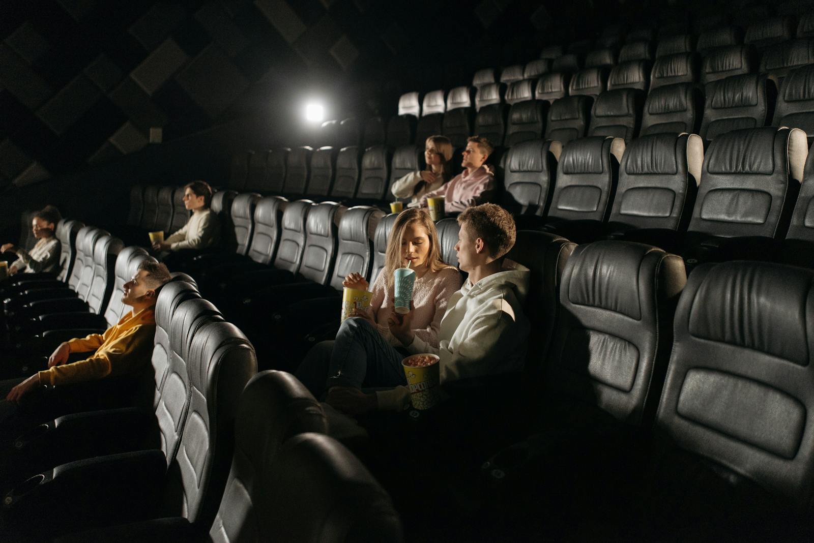 Couple sharing popcorn in a dimly lit movie theater with other viewers scattered.