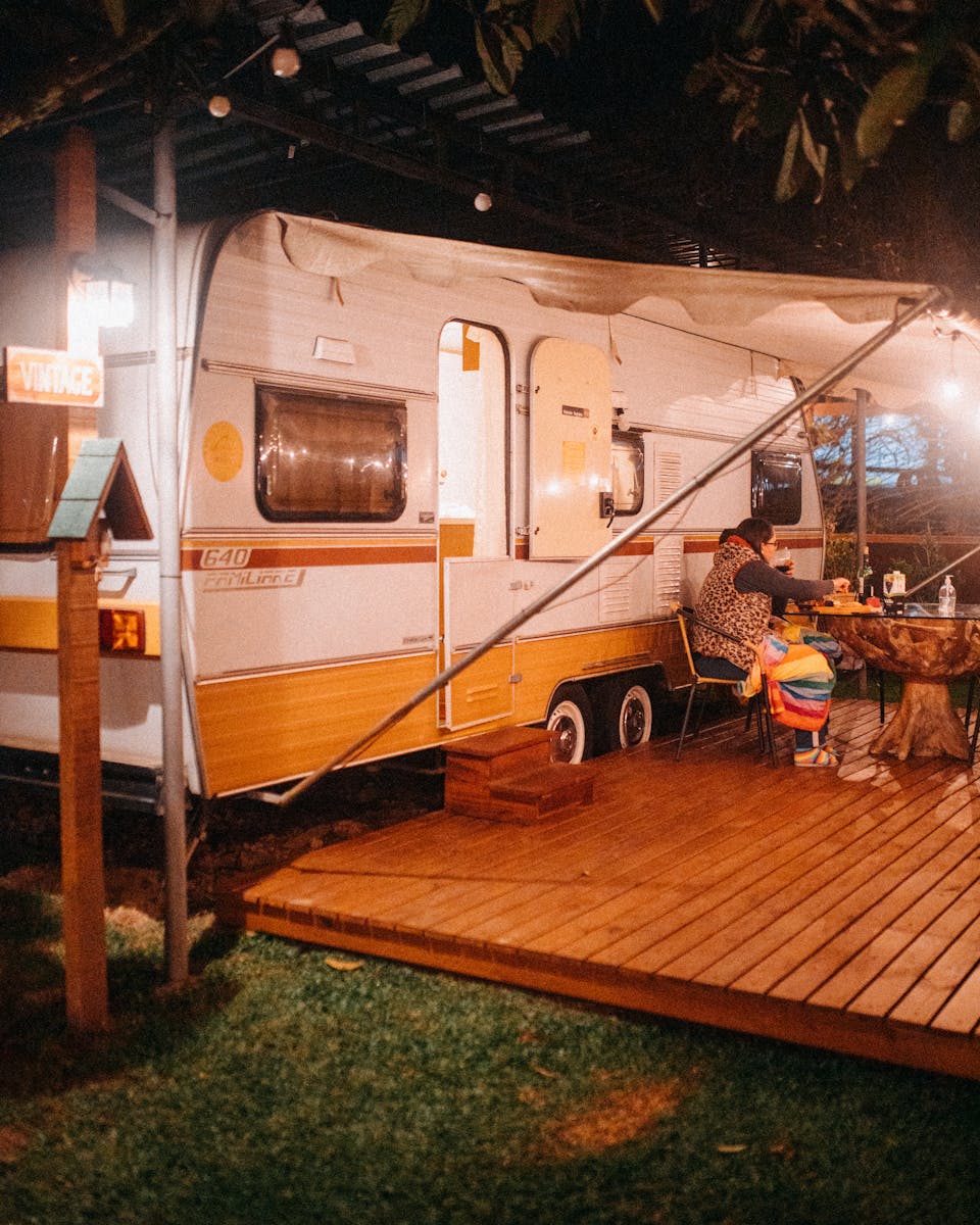 Unrecognizable female in warm clothes sitting at table placed on wooden platform near camping van at night glowing with garlands