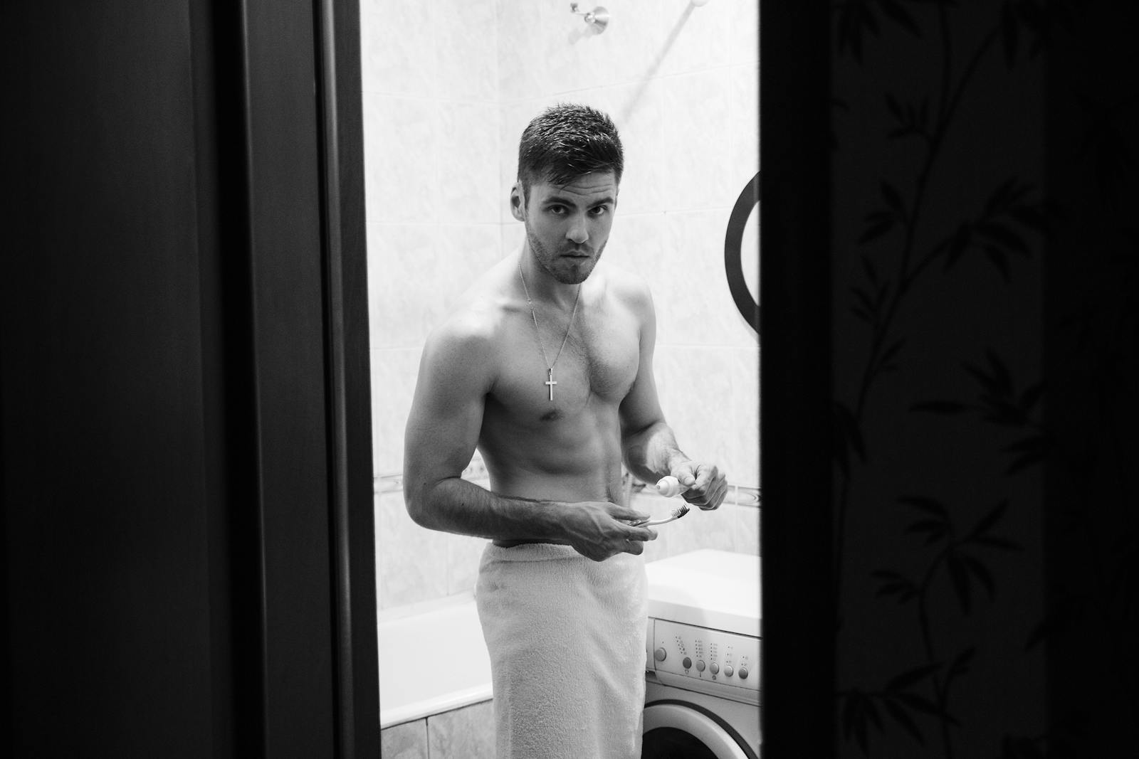 Black and white image of a shirtless man in a bathroom holding a toothbrush.
