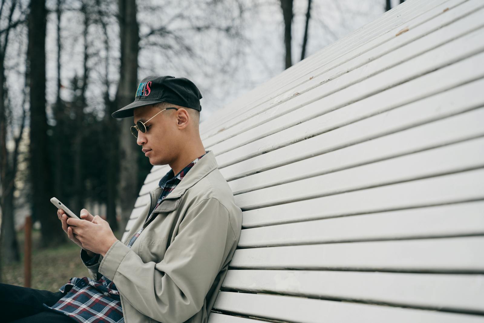 A young man in sunglasses and a cap texting on a smartphone while sitting on a park bench.