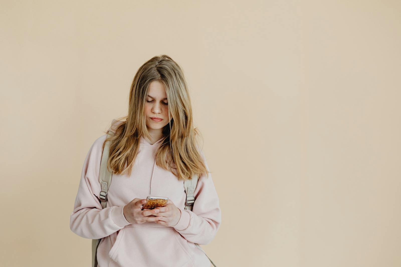 Teenage girl in hoodie and backpack checking smartphone against beige background.