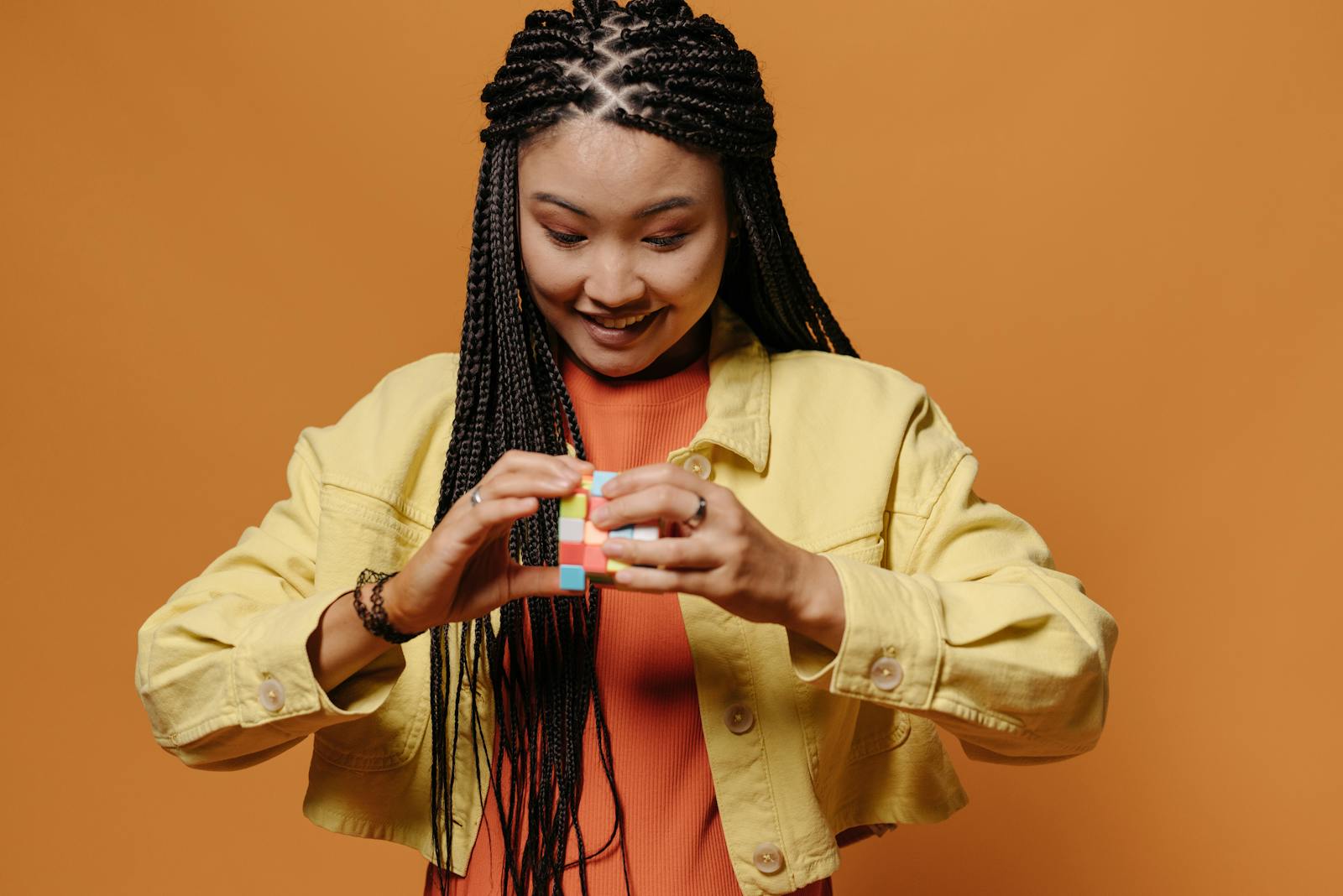 Portrait of a woman in a yellow jacket solving a Rubik's Cube against an orange background.