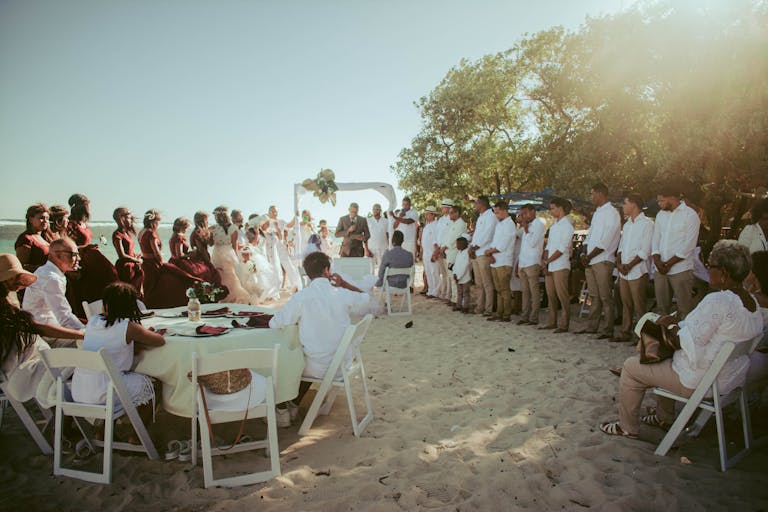 A beach wedding ceremony with a large group of guests in formal attire on a sunny day.