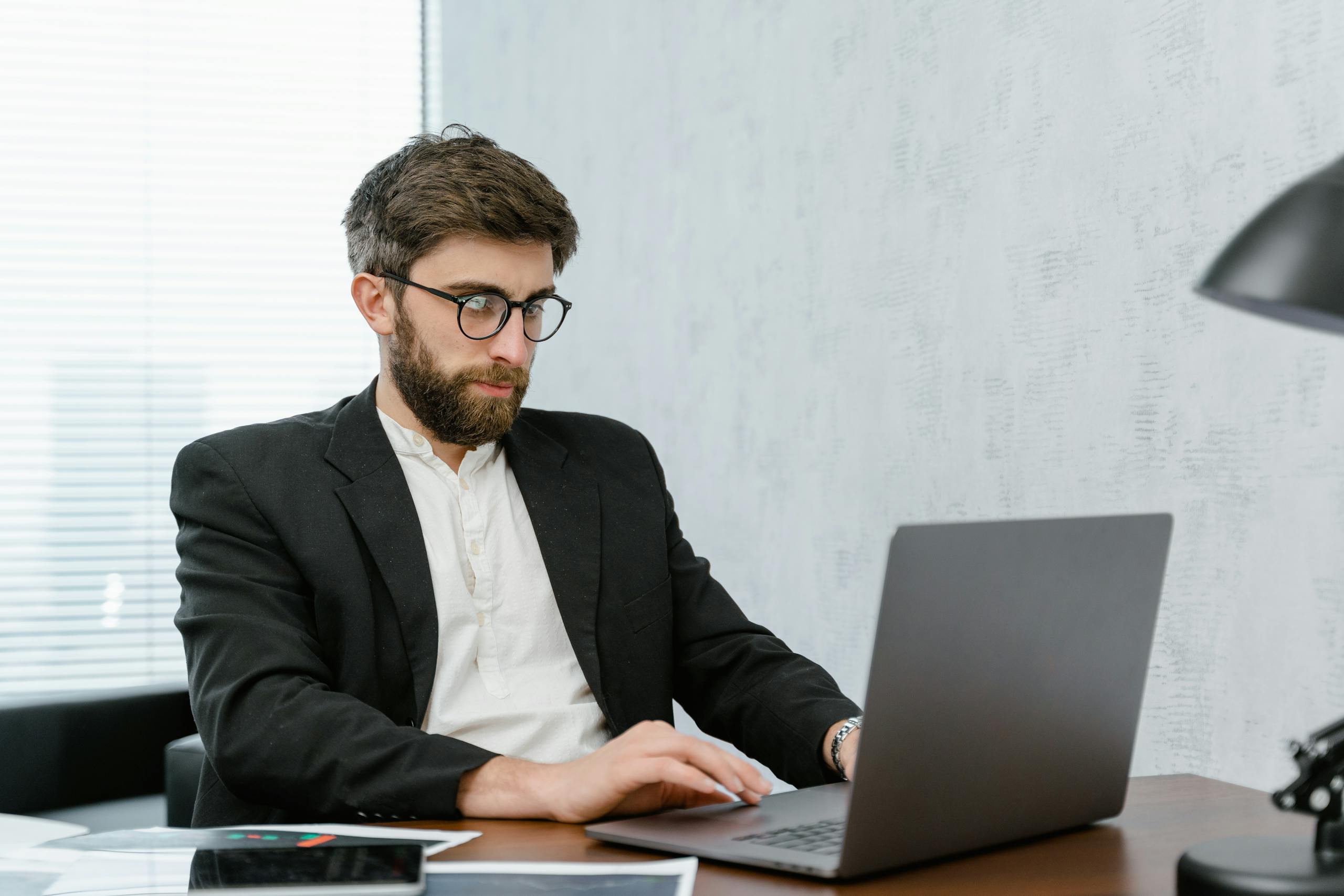 A bearded man in glasses and suit working on a laptop at an office desk.