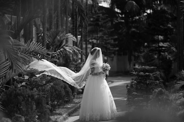A black and white photo of a bride with a flowing veil in a lush garden.