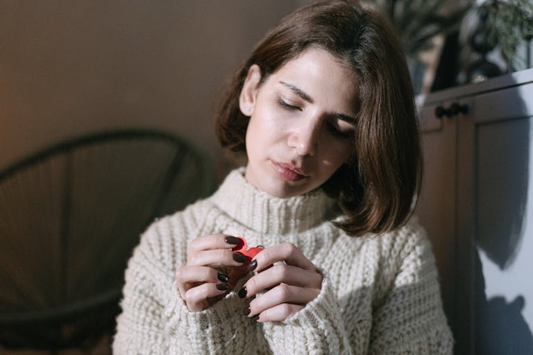 A brunette woman holding a red paper heart, looking thoughtful in a cozy indoor setting.
