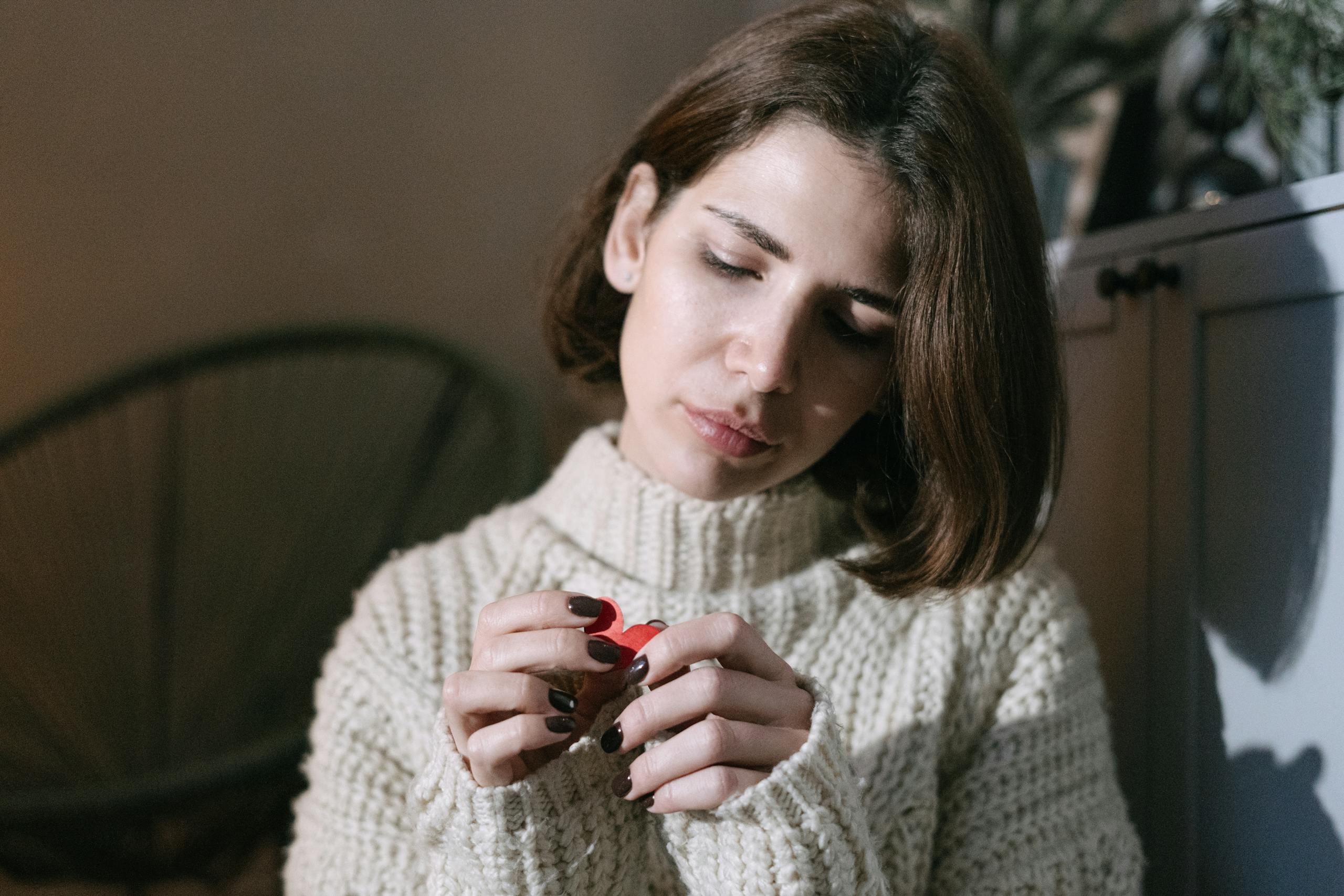 A brunette woman holding a red paper heart, looking thoughtful in a cozy indoor setting.