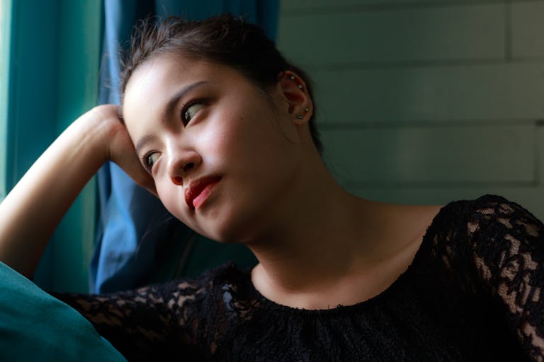 A contemplative woman in a lace dress gazes pensively by the window indoors.