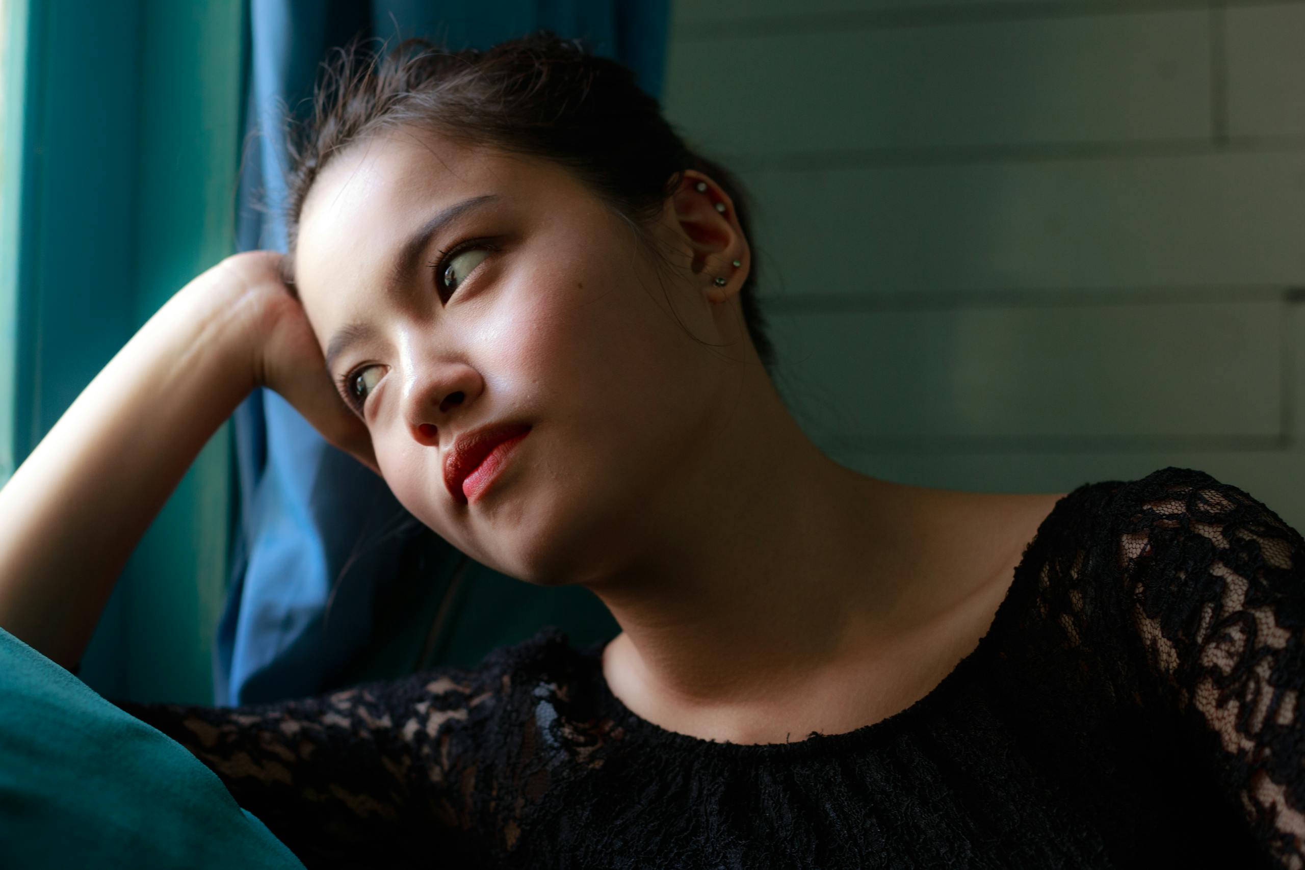 A contemplative woman in a lace dress gazes pensively by the window indoors.