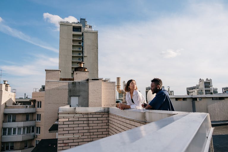 A couple chatting on a rooftop with a city skyline in the background under a clear blue sky.