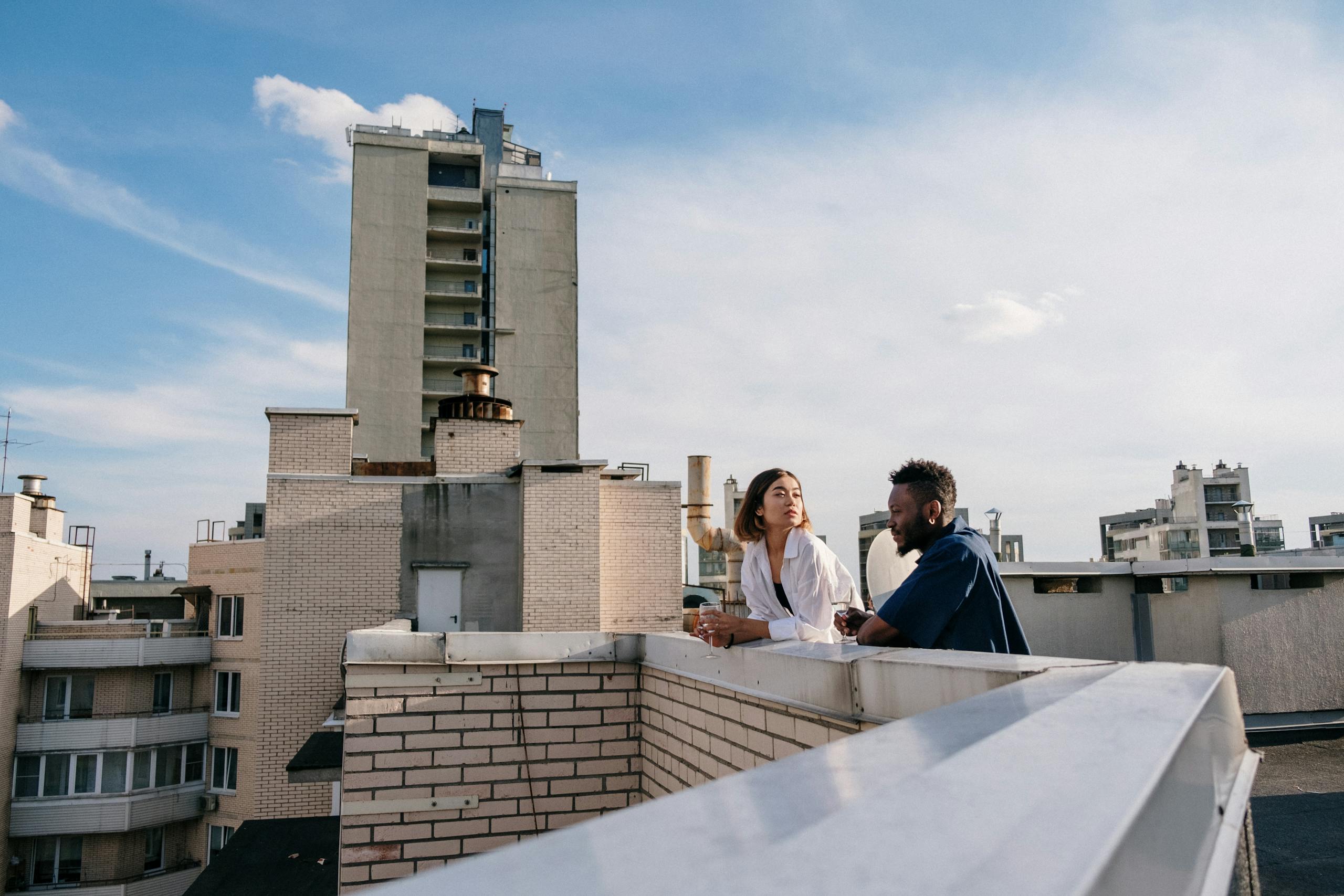 A couple chatting on a rooftop with a city skyline in the background under a clear blue sky.