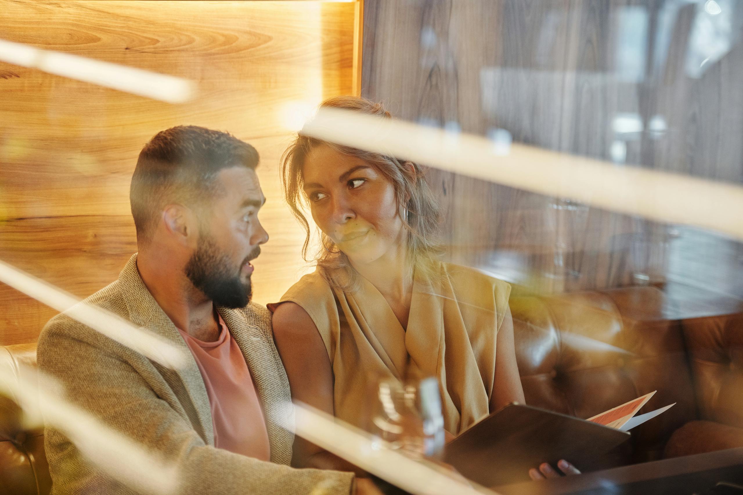 A couple enjoys a warm conversation in a cozy restaurant, viewed through a glass panel.