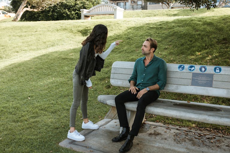 A couple in a heated argument while sitting and standing in a sunny park setting.