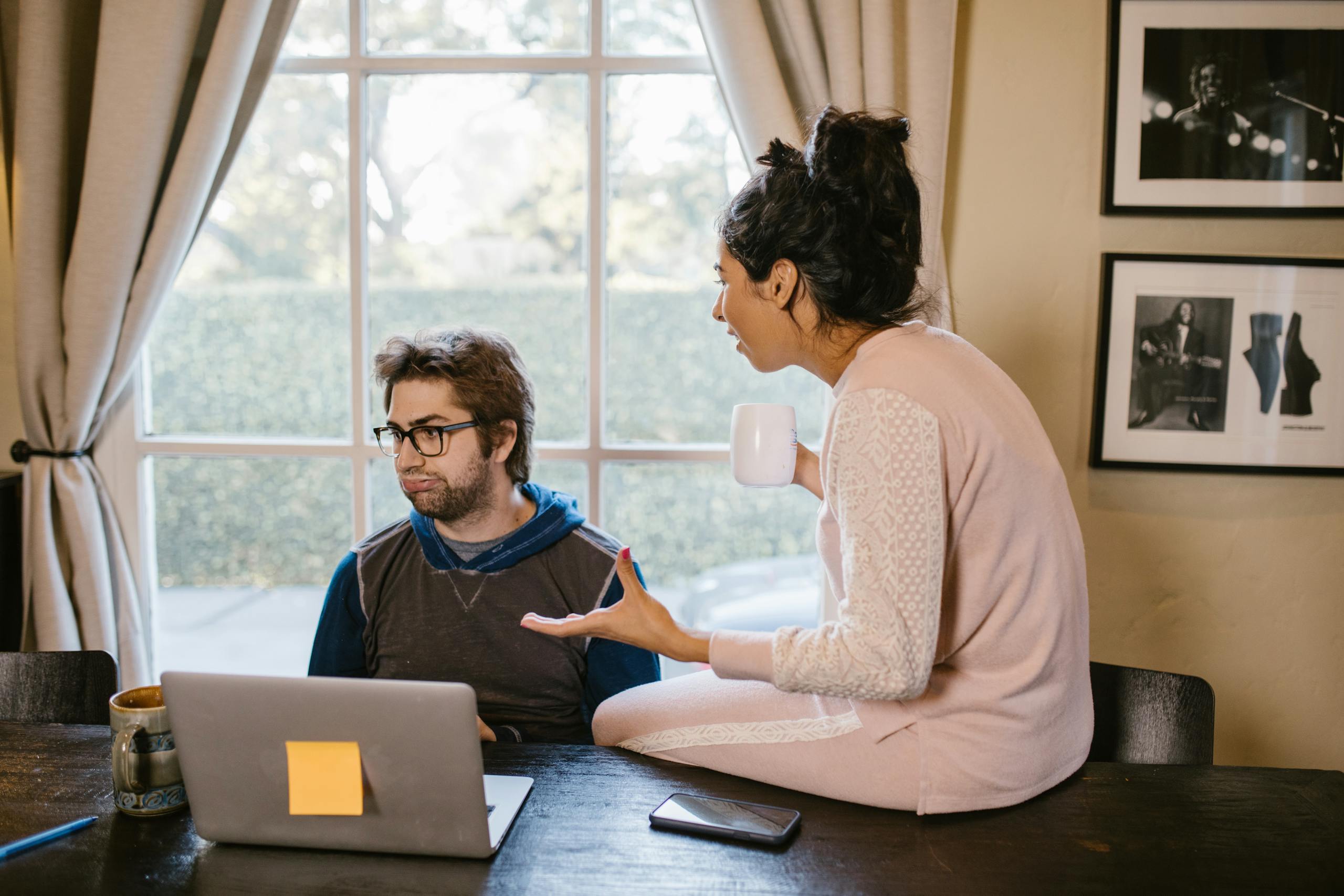 A couple sits at a table having a discussion, while working on a laptop indoors.