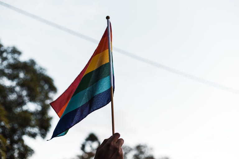 A hand holding a waving rainbow pride flag outdoors, symbolizing LGBTQ+ pride and unity.