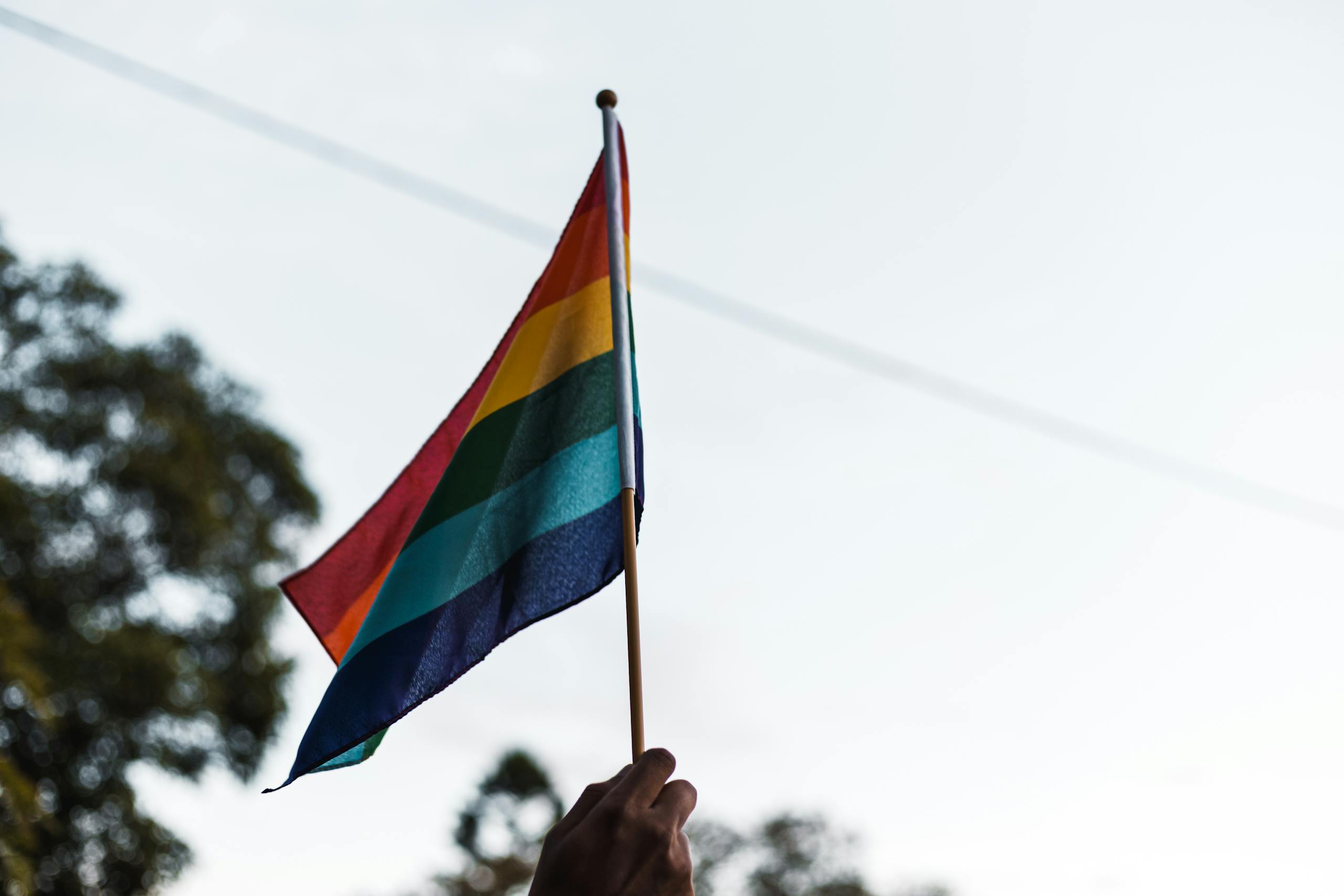 A hand holding a waving rainbow pride flag outdoors, symbolizing LGBTQ+ pride and unity.