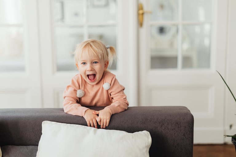 A joyful young girl with blonde hair laughing behind a couch indoors.