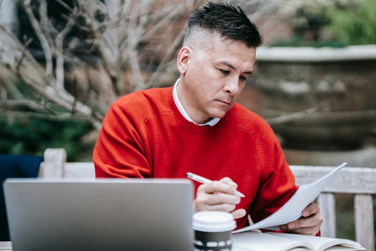 A man in a red sweater concentrating on paperwork while working remotely with a laptop outdoors.