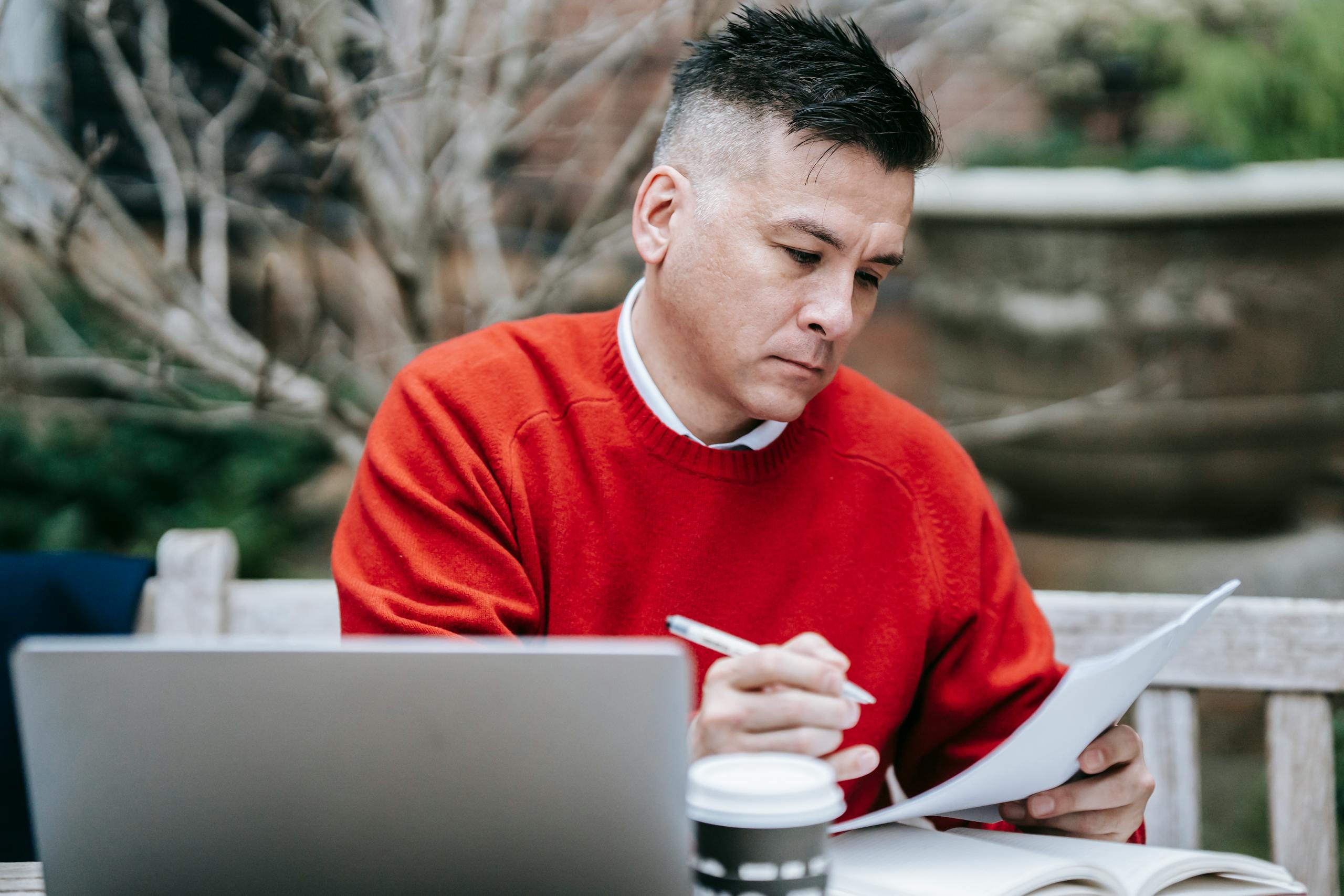 A man in a red sweater concentrating on paperwork while working remotely with a laptop outdoors.